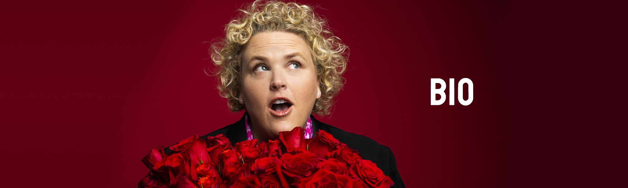 Comedian Fortune Feimster holding a large bouquet of red roses in front of a dark red background.
