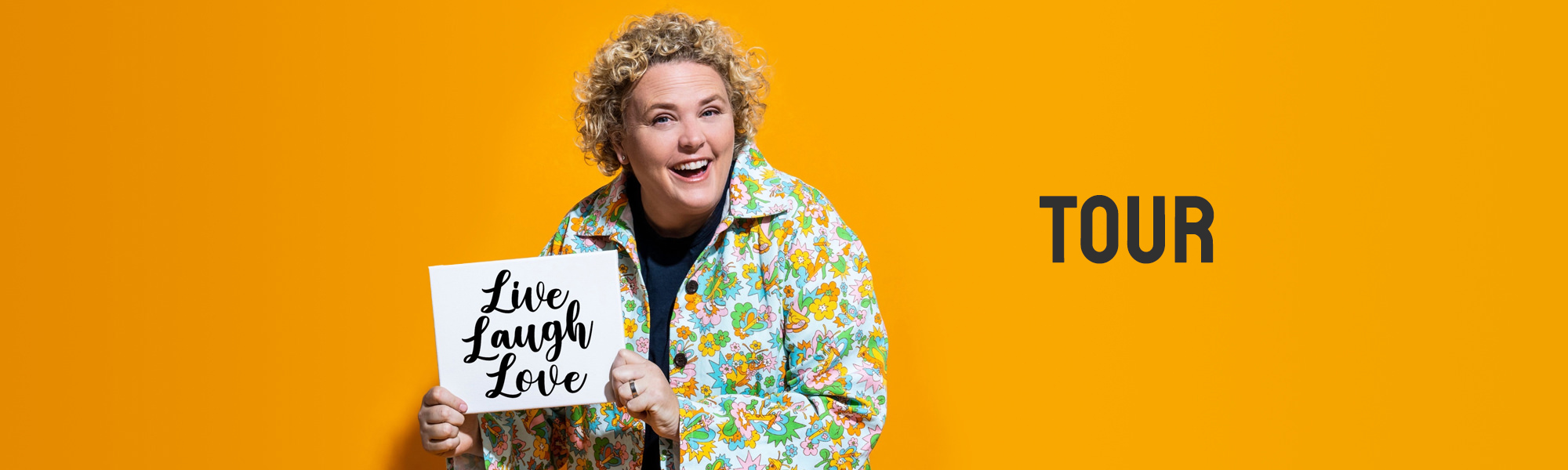 Comedian Fortune Feimster holding a sign that reads, 'Live Laugh Love' in front of a yellow background.