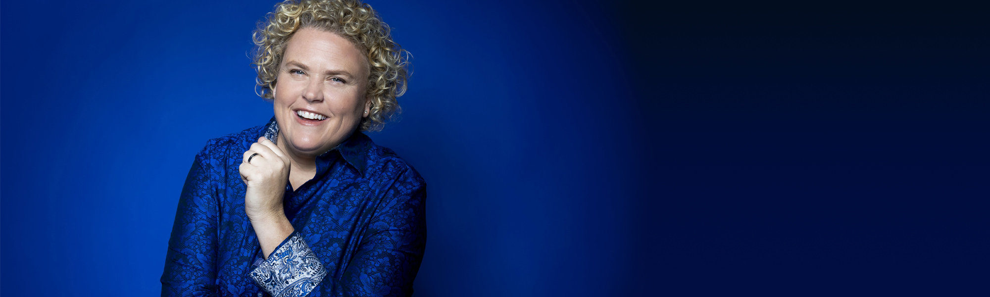 Comedian Fortune Feimster in front of a vivid blue background wearing a blue paisley shirt.
